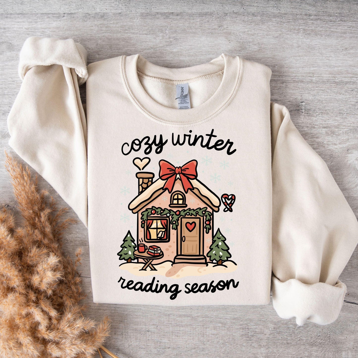 Flat lay of cream sweatshirt with cozy winter reading season gingerbread cottage print styled with warm textured blankets.