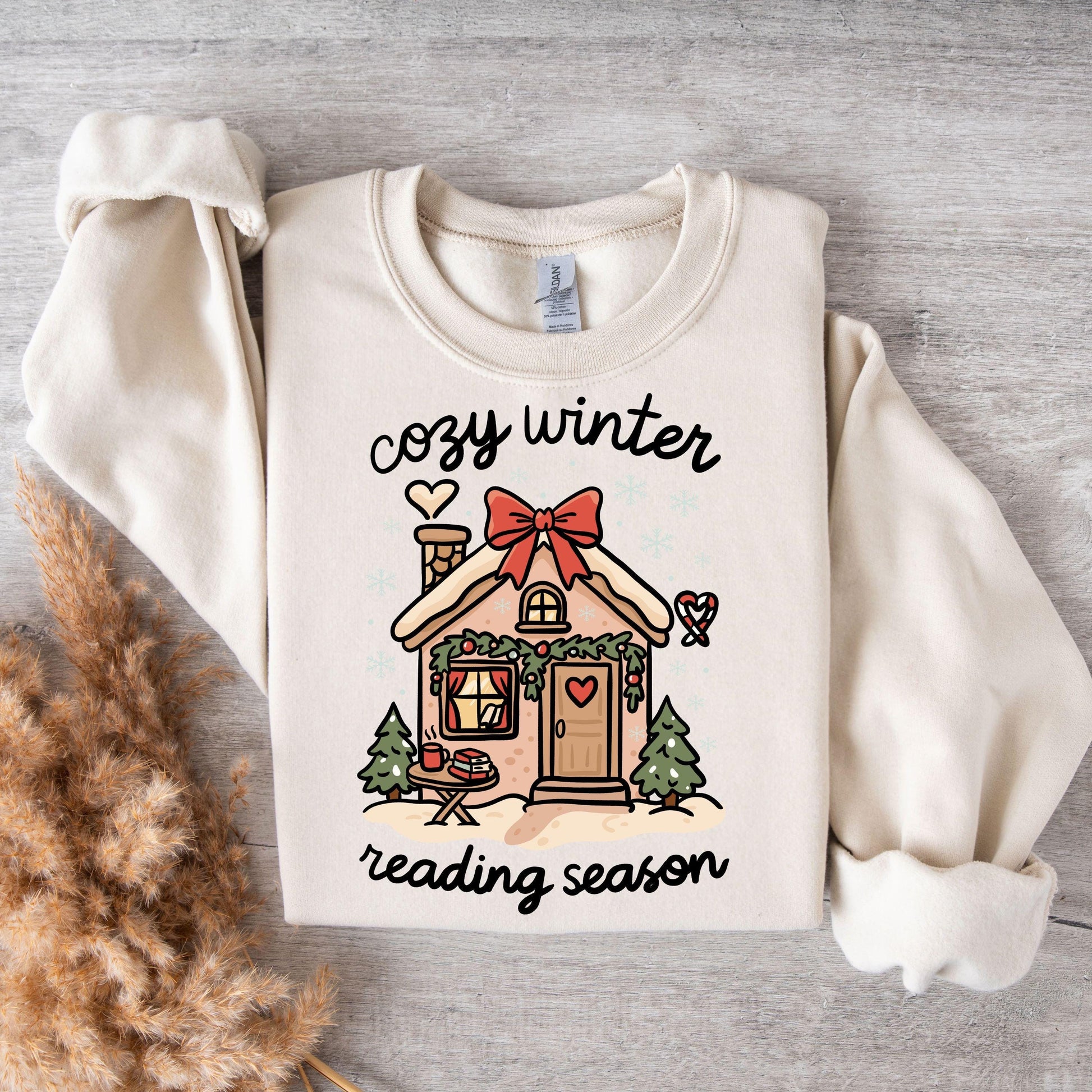 Flat lay of cream sweatshirt with cozy winter reading season gingerbread cottage print styled with warm textured blankets.