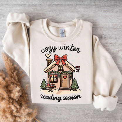 Flat lay of cream sweatshirt with cozy winter reading season gingerbread cottage print styled with warm textured blankets.