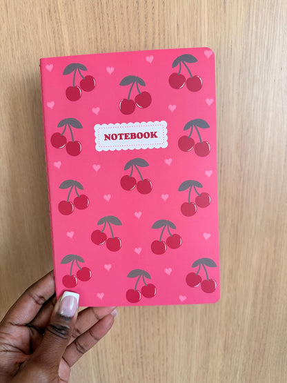 Pink notebook with a repeating cherry pattern and small heart accents, featuring a centered “Notebook” label on the cover.