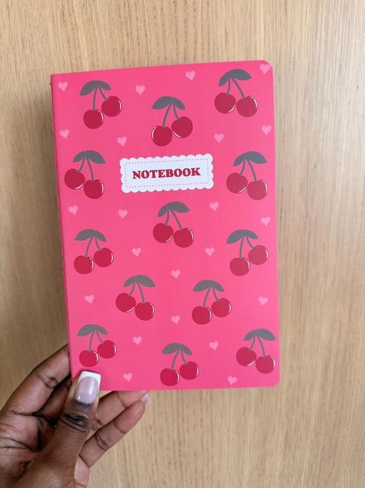 Pink notebook with a repeating cherry pattern and small heart accents, featuring a centered “Notebook” label on the cover.