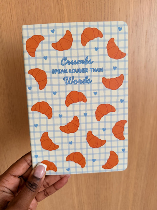 Croissant-themed notebook with a blue grid background and the phrase “Crumbs speak louder than words” on the cover, featuring illustrated croissants.